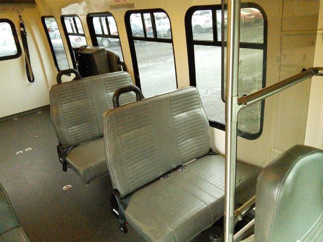Bus interior with gray vinyl seats arranged in rows, windows along sides, metal support poles, and a clean gray floor.