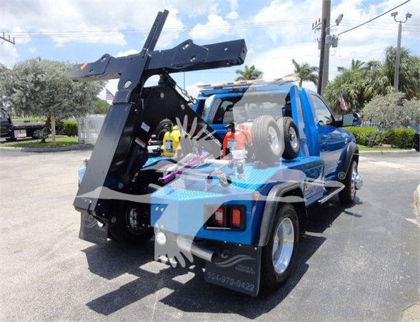 Tow truck with a blue cab and a heavy-duty hydraulic wheel-lift hoist in the rear, mounted on a flatbed. Visible text on the rear includes phone numbers and branding stickers.