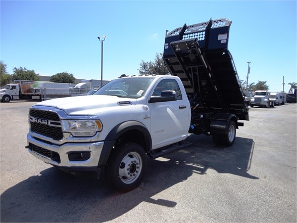 Ram 5500 HD dump truck in white with a raised black dump bed. Front grille shows RAM badge. Side shows 6.7L Cummins badge? Not legible. Industrial lot with other trucks.