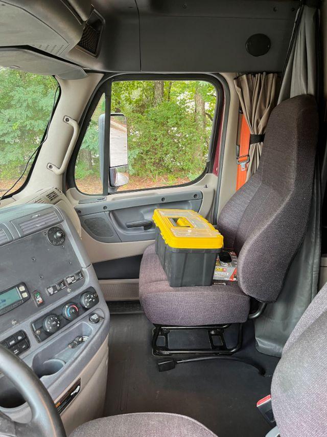 Cab interior of a 2015 Freightliner Cascadia truck showing dash controls, steering wheel, and passenger seat with a yellow and black toolbox on it.
