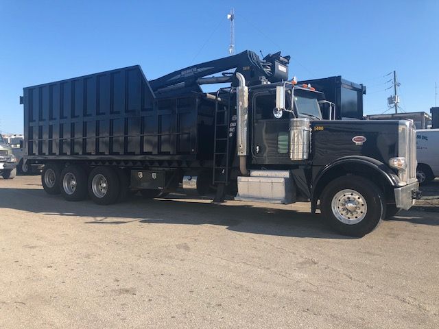 Peterbilt 388 dump truck with a grapple crane, black exterior, long dump bed, chrome bumper, dual rear axles, and a crane arm mounted behind the cab.