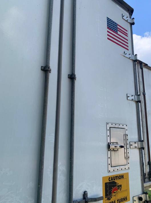 Trailer rear door with American flag sticker and warning label, vertical door hardware and latch on light gray surface.
