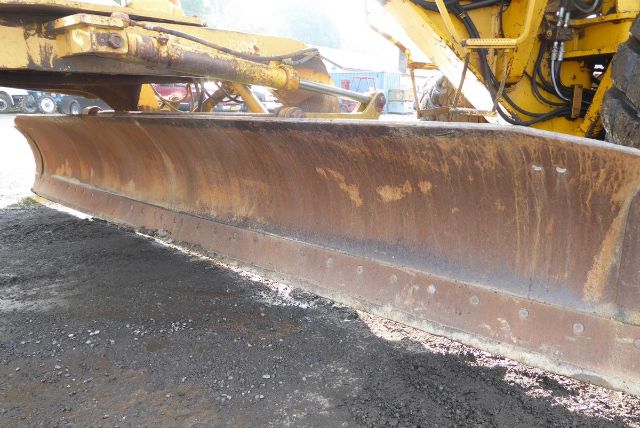 Caterpillar 16G grader blade with hydraulic arms and rusty edge, wide scraping surface, undercarriage not visible.