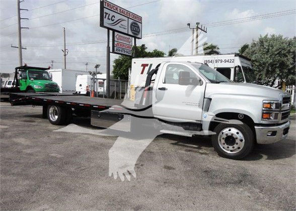 Flatbed tow truck with white cab and long black flatbed. Visible text on sign: 'TRUCK SALES CENTER TLC' canopy in background. No other legible model or spec text clearly readable.