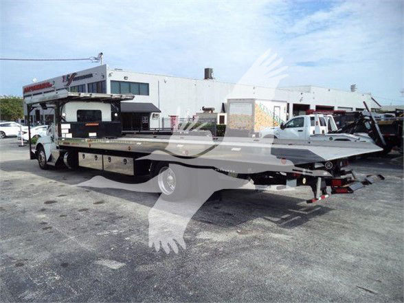 Kenworth T370 tow truck with flatbed; visible side cabinets and equipment; company logos appear on the truck and building in background; no legible fine print close-up.