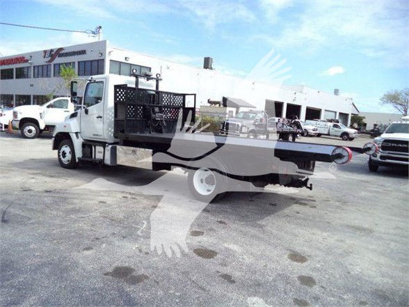 2017 Hino 258LP hooklift truck in a dealership lot, side view with flatbed and hooklift mechanism visible, white cab, dealership buildings in background.