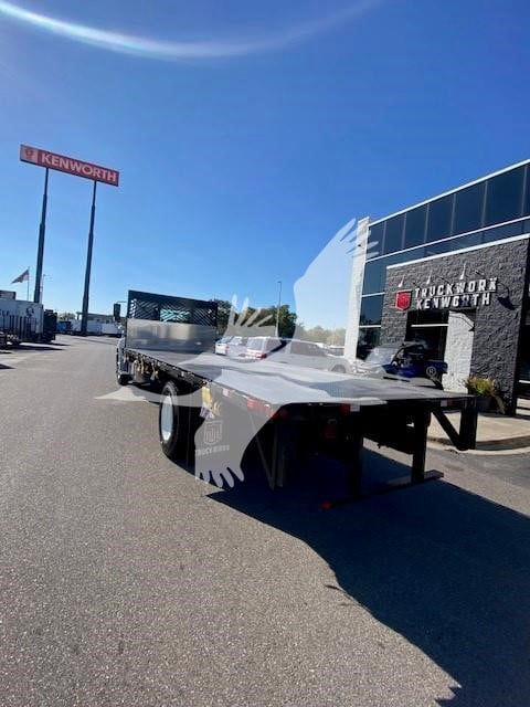 Flatbed truck chassis in a dealership lot with Hino 2025 model, VIN 5PVNJ7AV1S5T58302, stock HI1785, new, Kenworth/Truck World signage in background.