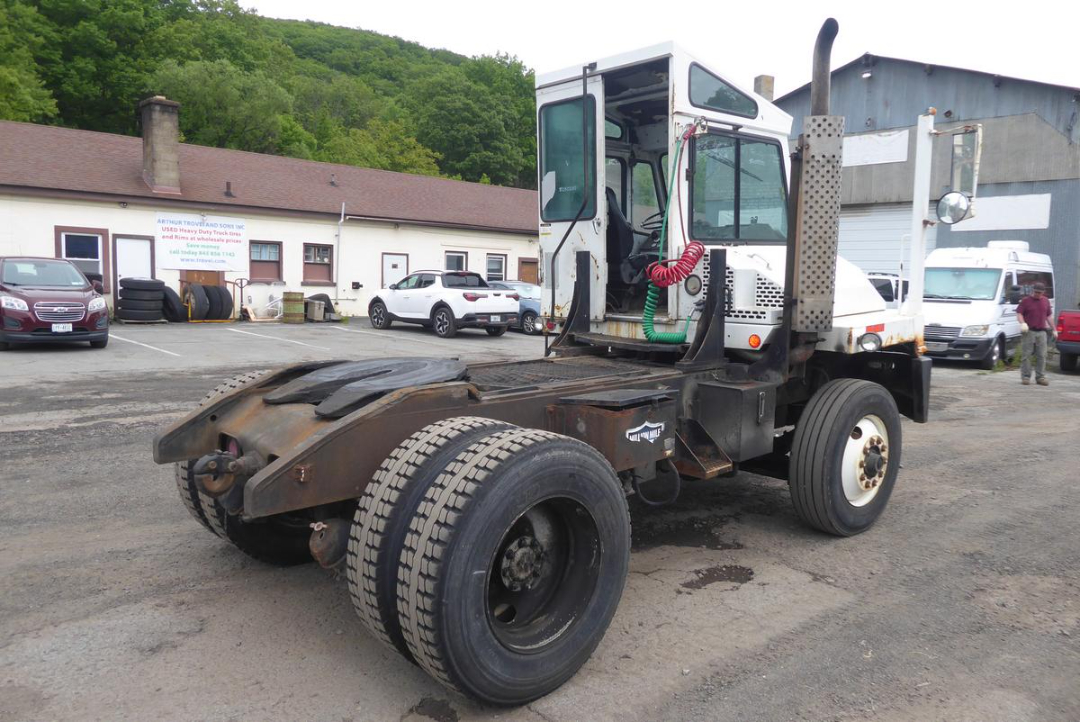 Truck tractor chassis with cab, rear dual rear wheels, and fifth wheel plate showing a rusty frame and fuel lines; white cab with green and red hoses.