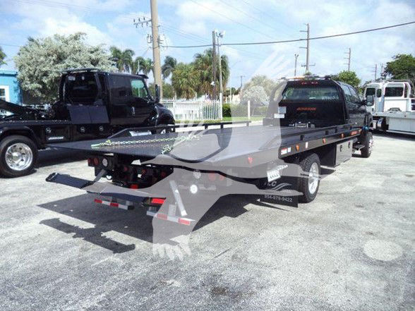 Tow truck flatbed trailer with ramp in a dealership lot, International CV visible in listing. Includes chain tie-downs and wheel chocks.
