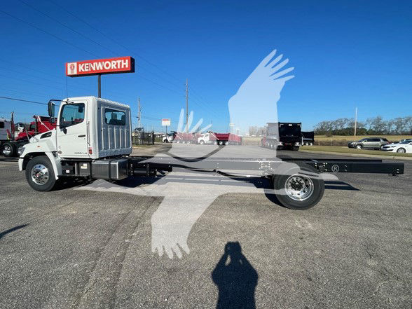 2025 Hino L6 cab and chassis truck with long wheelbase and flatbed; cab in front, chassis extending to rear; dealership lot with Kenworth sign in background.