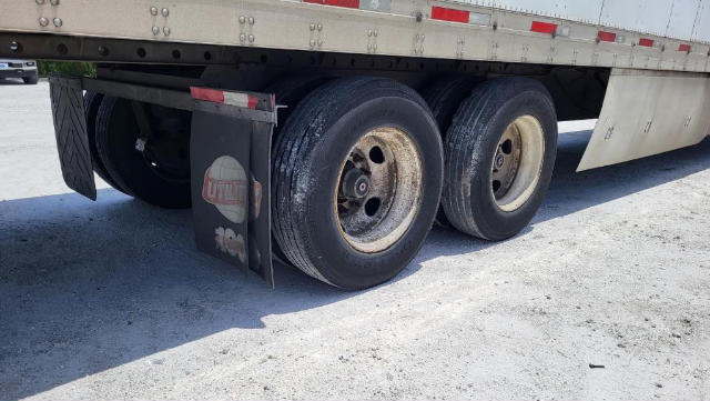 Dual rear axles on a utility trailer with black tires, steel rims, and mud flaps showing a faded logo.