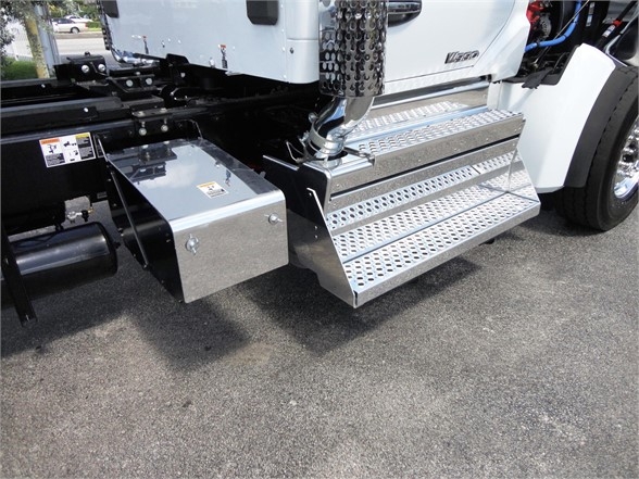 Truck with metal side steps and a diamond plate running board. Visible components include a storage box on the frame, a chrome exhaust stack cover, and a portion of the cab.