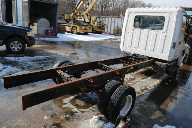 Cab and chassis truck frame with dual rear wheels and white cab, snow on ground, yard with equipment in background.