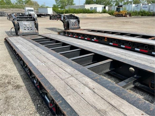 Lowboy trailer deck with steel beams and gooseneck legs visible; industrial surroundings