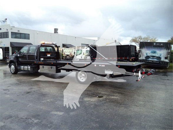 Flatbed tow truck with orange beacon and black flatbed; text on cab windows and trailer not fully legible.