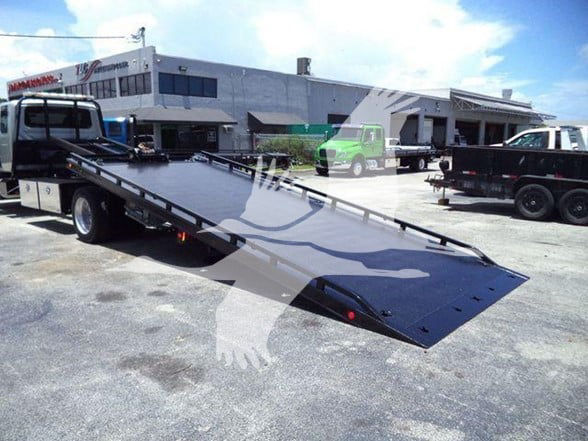 Tow truck flatbed with loading ramp extended in a dealership lot.