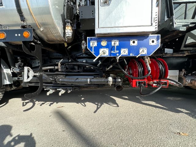 Underbelly of a 2013 International 7500 vacuum truck showing hydraulic lines, a blue control panel with gauges, red hose reel, and black chassis components.