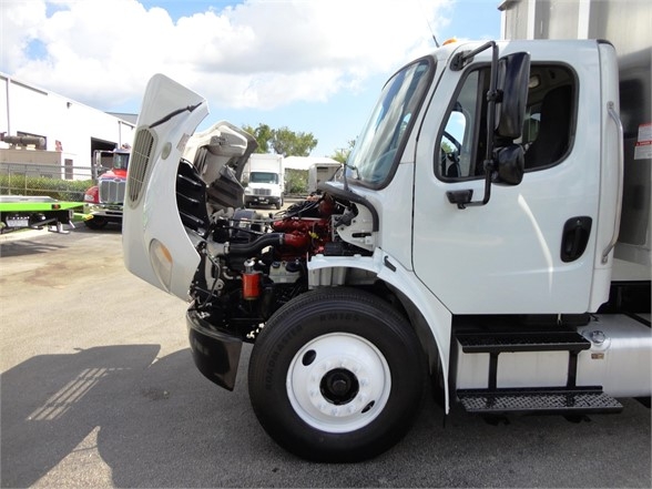 Truck engine bay visible with hood open on Freightliner Business Class M2 106; white cab, exposed red and black engine components, front wheel and step.