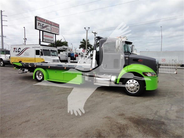 A 2022 Peterbilt 337 tow truck with a green and gray cab, flatbed, and chrome accents parked in lot; TLC and other trucks visible in background.