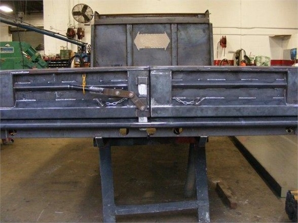 Dump body truck bed showing metal tailgate and lock chains, mounted on a stand in a workshop.