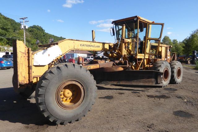 Caterpillar 16G motor grader in a yard showing large front wheel, boom blade, and operator cab with yellow paint and surface grime.