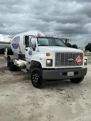 Truck with a cylindrical tank mounted on a chassis, white cab, large front grille, hazard diamond placard on the passenger side of the bumper, and a logo on the tank. Visible text includes a circular sign on the tank, side text near the doo