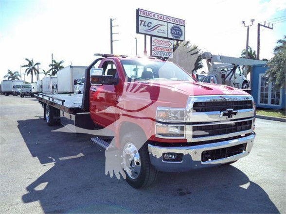 Flatbed tow truck with red cab and Chevrolet emblem; front grille; dealership sign in background; palm trees; blue sky.