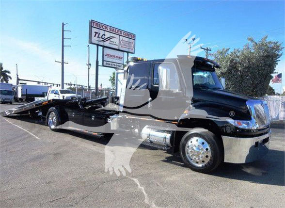 Tow truck showing International MV cab and a low-profile car carrier bed with reflective chrome wheels. Text visible: Truck Sales Center TLC sign in background; dealership lot.