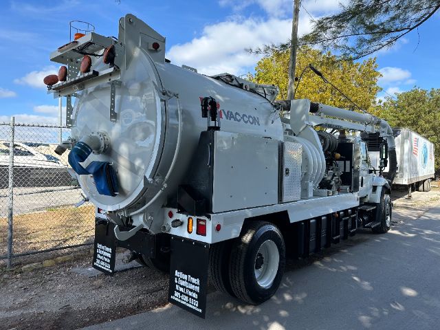 Vacuum truck with large cylindrical rear unit labeled VAC-CON on a flatbed truck, connected hose assembly, blue sky and trees in background.
