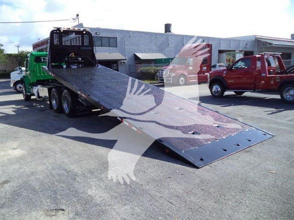 Freightliner Business Class M2 106 tow truck with a lowered flatbed/wrecker bed on a tilt ramp, green truck cab and red trucks in background.