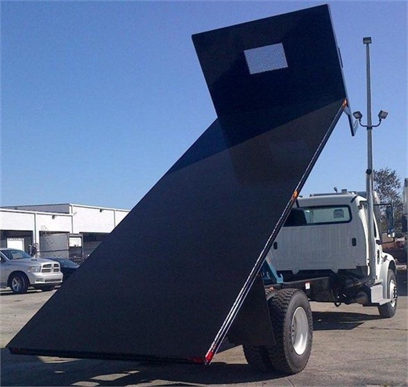 Flatbed dump truck with a raised black steel bed against a clear blue sky; cab and front wheels visible; site shows industrial area.