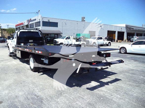 Tow truck transporter flatbed with extended long deck and hooks, rear view showing black flatbed and wheel wells, light gray building in background.
