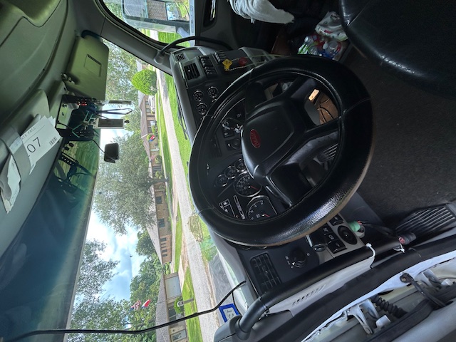 Truck cab interior with steering wheel and dashboard of a Peterbilt 579; visible gauges and hanging tags near the windshield.