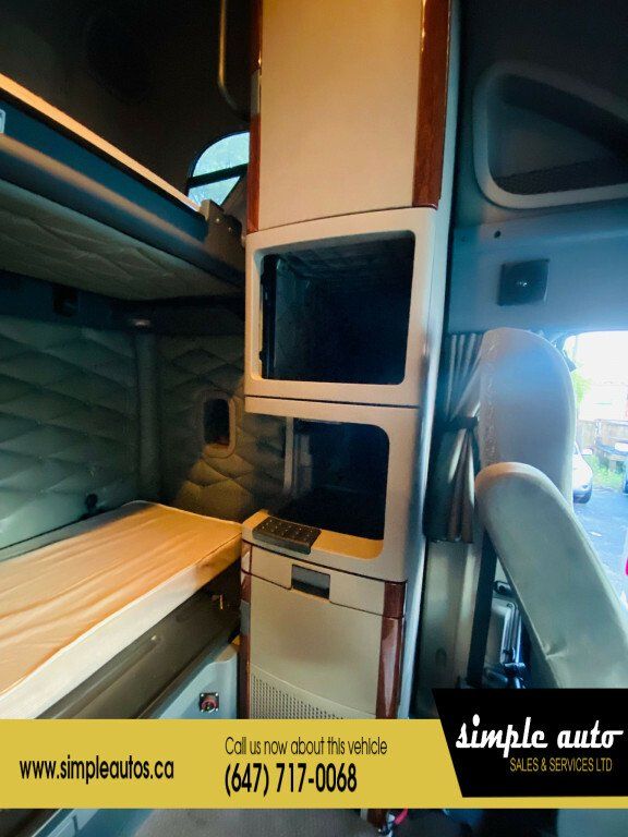 Cab interior of a 2012 Freightliner Cascadia showing upper bunk bed area, storage cabinets, and control panel; vertical wood trim, quilted green/gray upholstery.