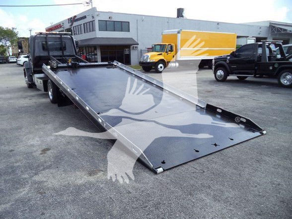 2014 Hino 258ALP tow truck with a long flatbed ramp extended, in a dealership lot. Visible are a black tow truck in background, a white building, and other trucks.