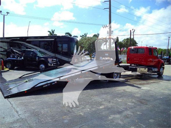Tow truck with a flatbed, MV607RED stock number visible in listing; red tow truck in background; clear sky with scattered clouds.