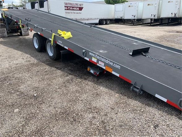 Flatbed trailer with a low-angle ramp, tandem axle, black paint, yellow safety latch, and reflective red/white strips.