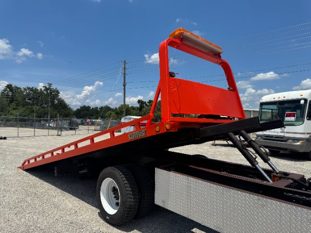 Tow truck flatbed street-angled view showing red flatbed with hydraulic ramps and a black frame, International-style truck bed mid-length, blue sky background.