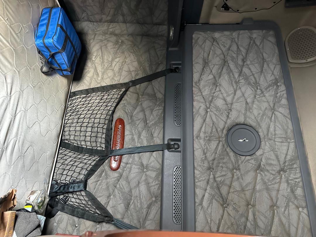 Freightliner Cascadia sleeper cab interior showing quilted mattress, tie-down nets, storage pouch, and circular fuel tank access lid in the floor with a small speaker on the wall.