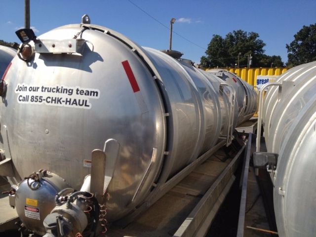 Vacuum tank trailer showing cylindrical stainless tank with decals and fittings; text on tank reading 'Join our trucking team Call 855-CHK-HAUL' and various valves; shiny metallic exterior; conveyor/rail structure beneath.