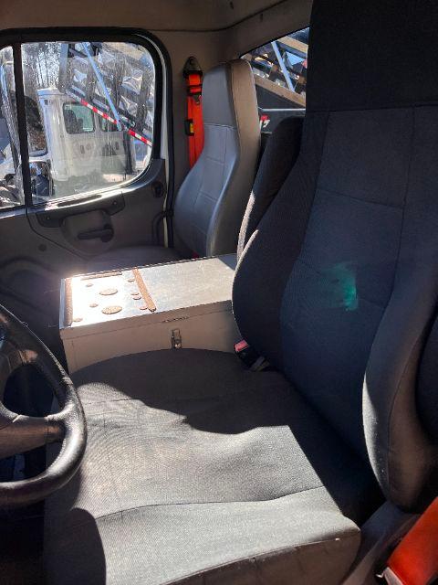 Cab interior of a 2015 Freightliner M2 truck showing driver's seat, passenger seat, steering wheel, and a metal center consul with cup holders and latch. Side window and door visible.