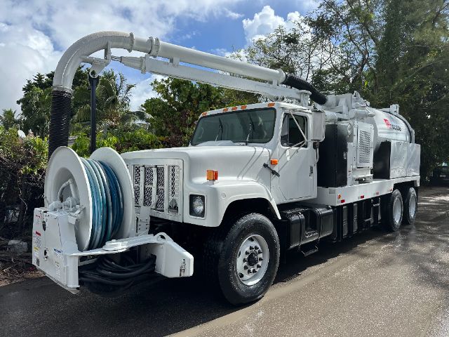 Vehicle vacuum truck showing front cab and hose reel with large hose on reel and long intake pipe on top; International truck with grey body.