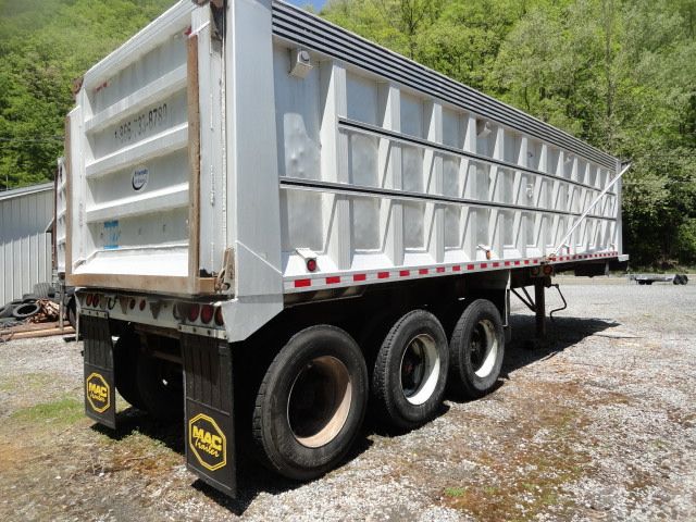 Tri-axle dump trailer with three axles and large metal side walls. Visible are MAC mud flaps, tire rims, and rear tailgate with faded markings.