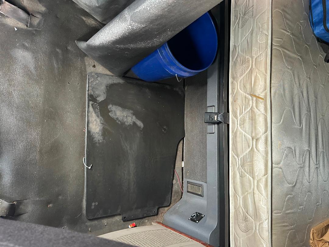 Truck cab interior showing a storage area with a blue bucket, storage mat, and mattress edge. View into cabin with plastic trim and control panels.