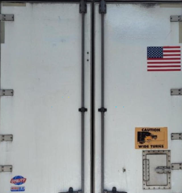Rear doors of a utility trailer with vertical dual doors, American flag sticker on right door, CAUTION WIDE TURNS sticker near bottom right, small round sticker bottom left, and a couple of latches.