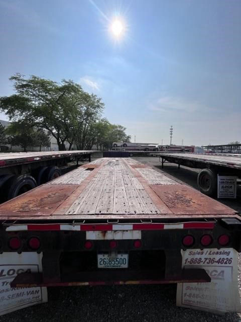 Flatbed trailer deck with wooden planks, rear view showing taillights and registration plate; Fontaine trailer lot in background under clear sky.