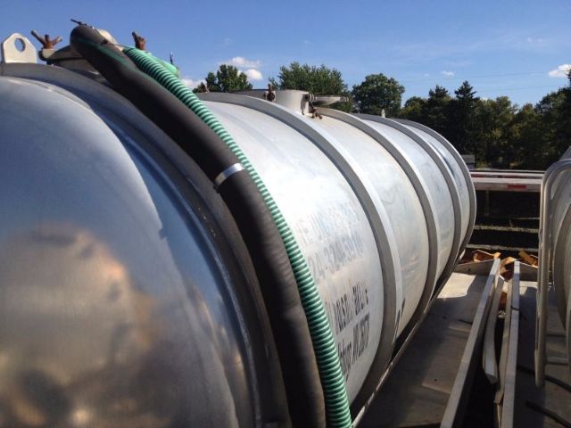 Vacuum tank trailer cylinder on a flatbed with hose attached and reflective surface.