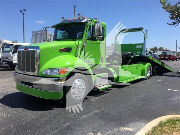 Green Peterbilt tow truck with hydraulic rollback bed, front chrome bumper, cab with large grille, and extended flatbed in green color.