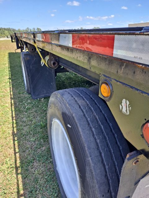 Flatbed trailer side rail with spare tire, warning lights, and reflective tape; visible weathered frame and wheel on top of bed.