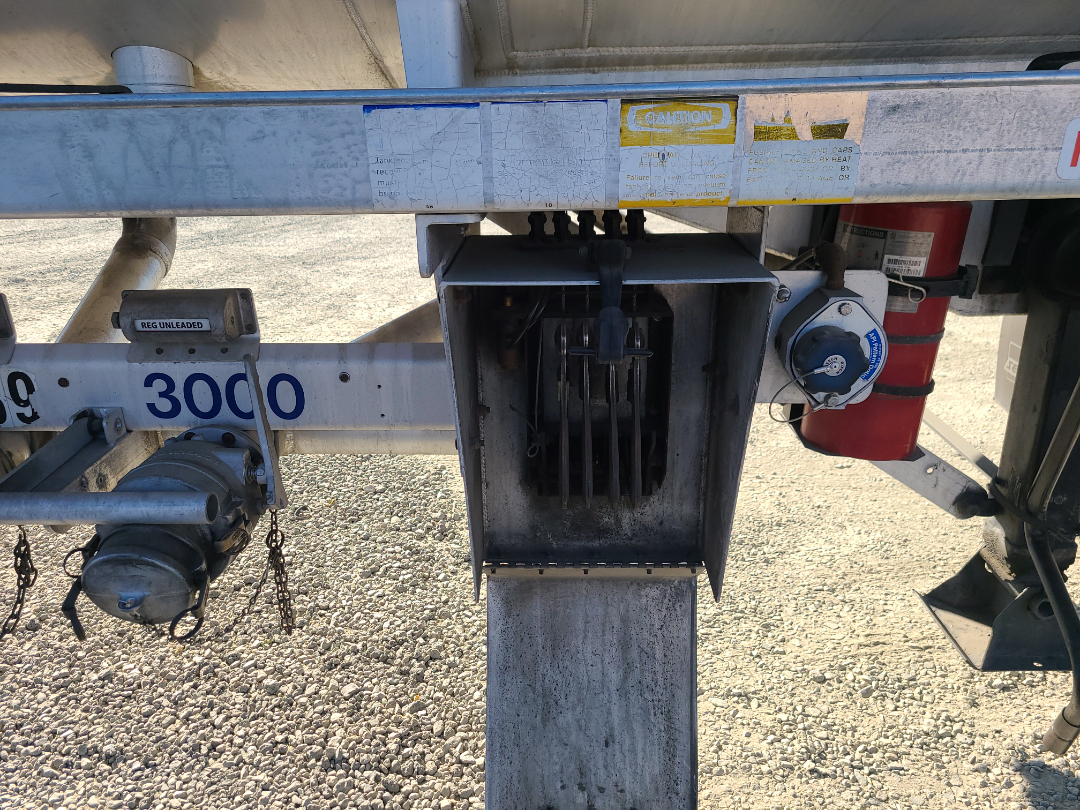 Trailer hitch area showing Beall semi trailer with central fuel/vent box and safety equipment. Visible labels include caution stickers, a red fire extinguisher, and a gray electrical/control box inside a weathered metal housing.